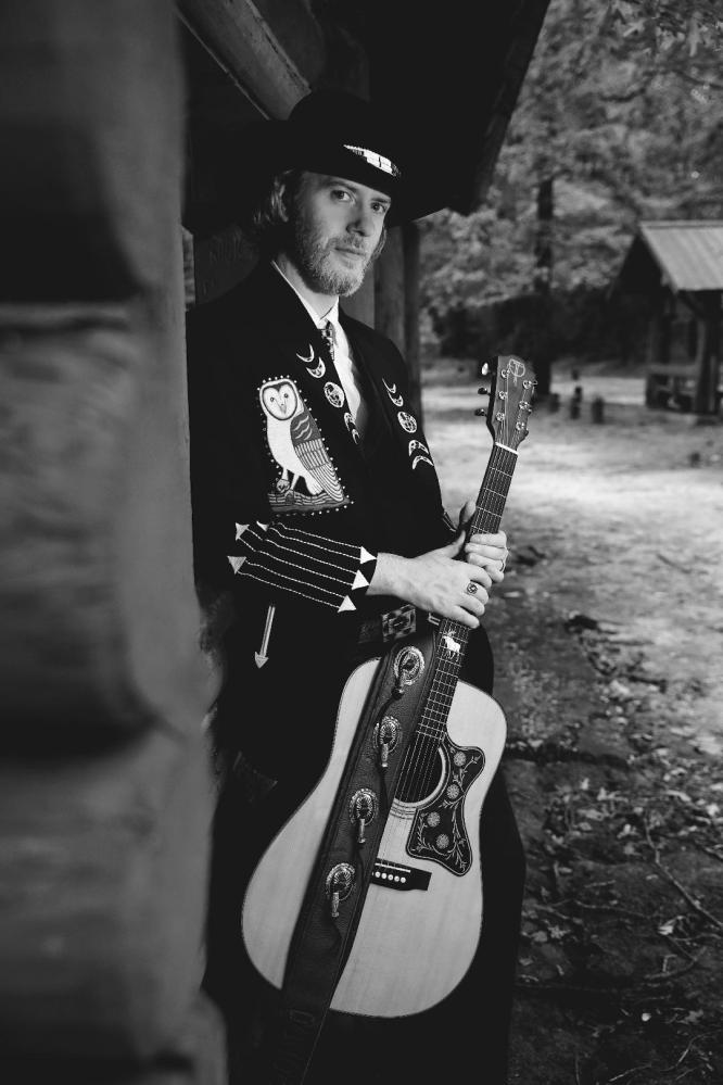 Man in hat with guitar leans against wooden structure in forest setting, wearing a suit with owl and moon designs.