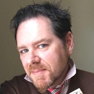 Man with short hair and beard wearing a dark sweater and striped shirt, looking into the camera.