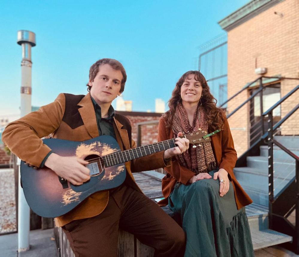 Two people sitting; one plays a guitar on a rooftop with blue sky.