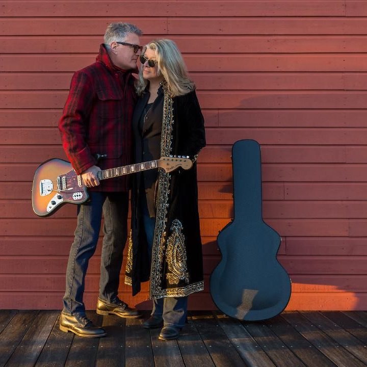 Couple hugging with a guitar against a red wall, guitar case on the ground.