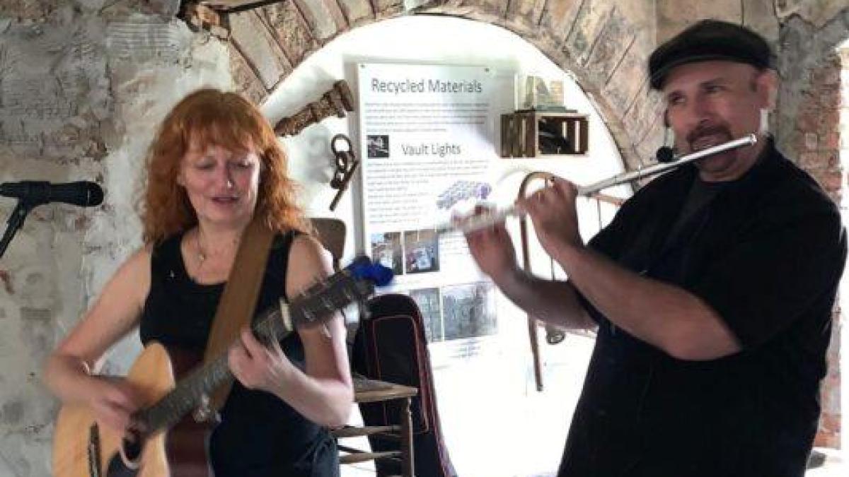 Woman playing guitar and man playing flute in rustic room with stone walls.