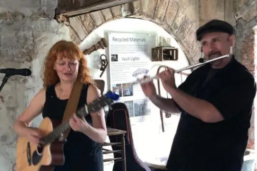 Woman playing guitar and man playing flute in rustic room with stone walls.