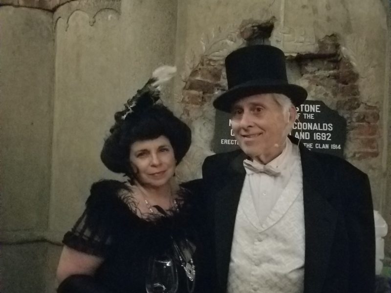 A couple in vintage formal attire by a rustic stone wall with arches.