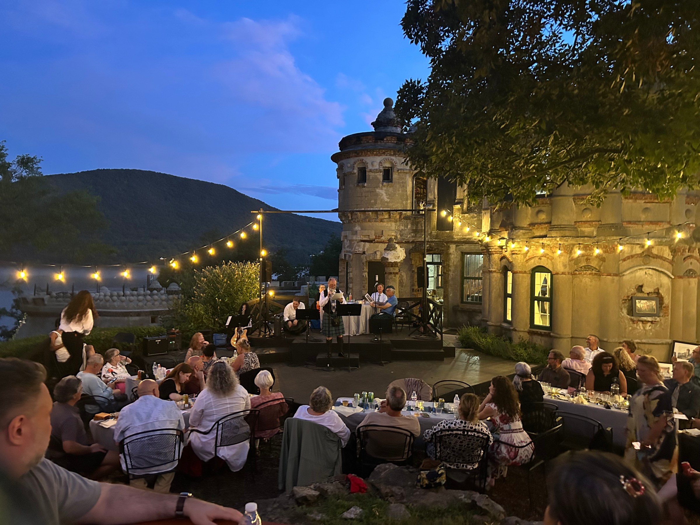 Outdoor event with people dining, string lights, and a band playing near a castle-like building at dusk.