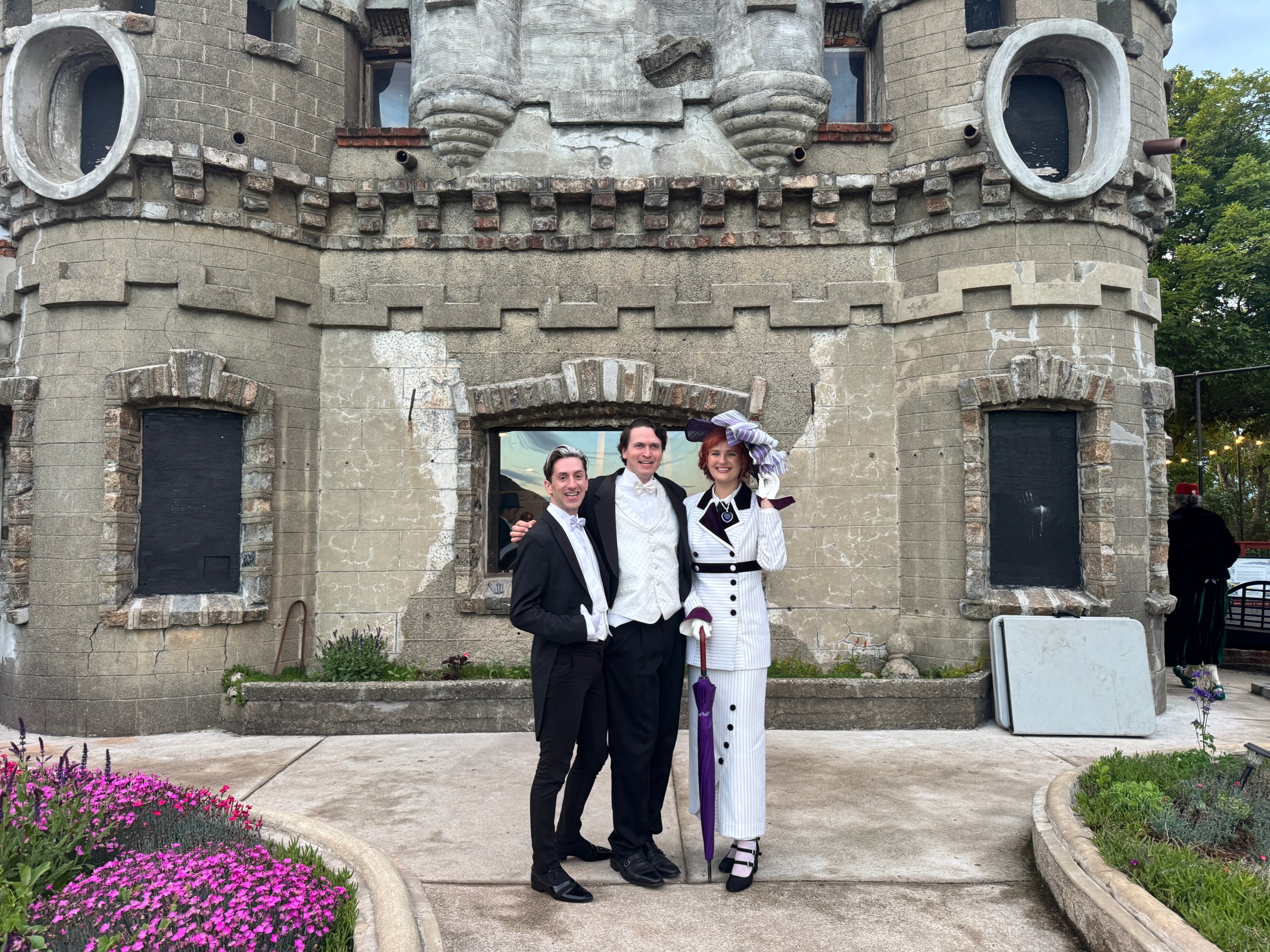 Three people in vintage attire pose in front of a castle-like building with circular windows.