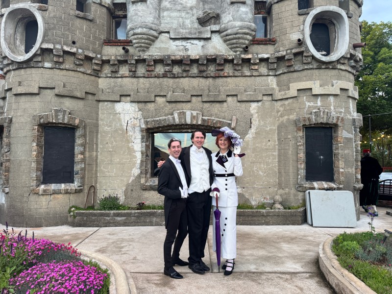 Three people in vintage attire pose in front of a castle-like building with circular windows.