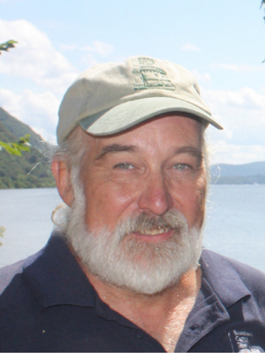 Man in cap standing by a lake with mountains in background.