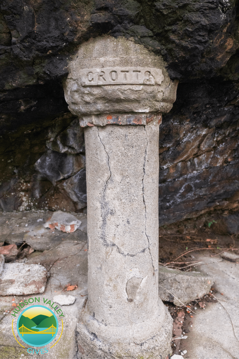Weathered stone column with 'GROTTA' inscribed, surrounded by rocks, and cracked at the base.