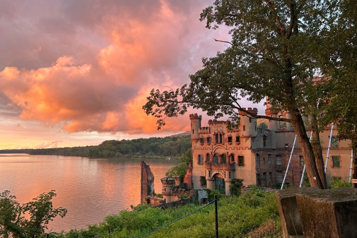 Historic castle by a lake at sunset with vibrant orange clouds and lush greenery.